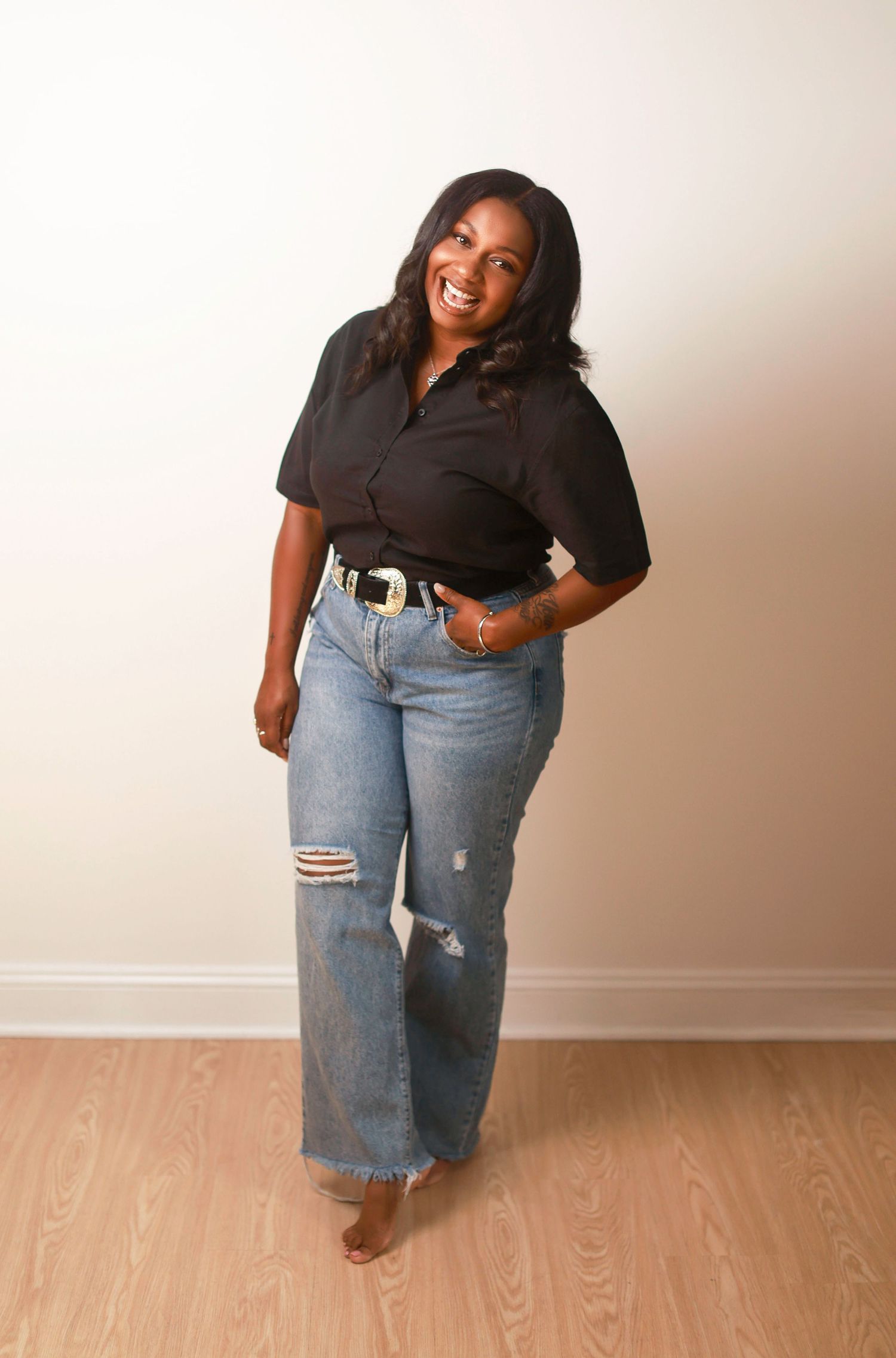 A person wearing a black top and distressed blue jeans poses playfully against a beige wall in a photo series.