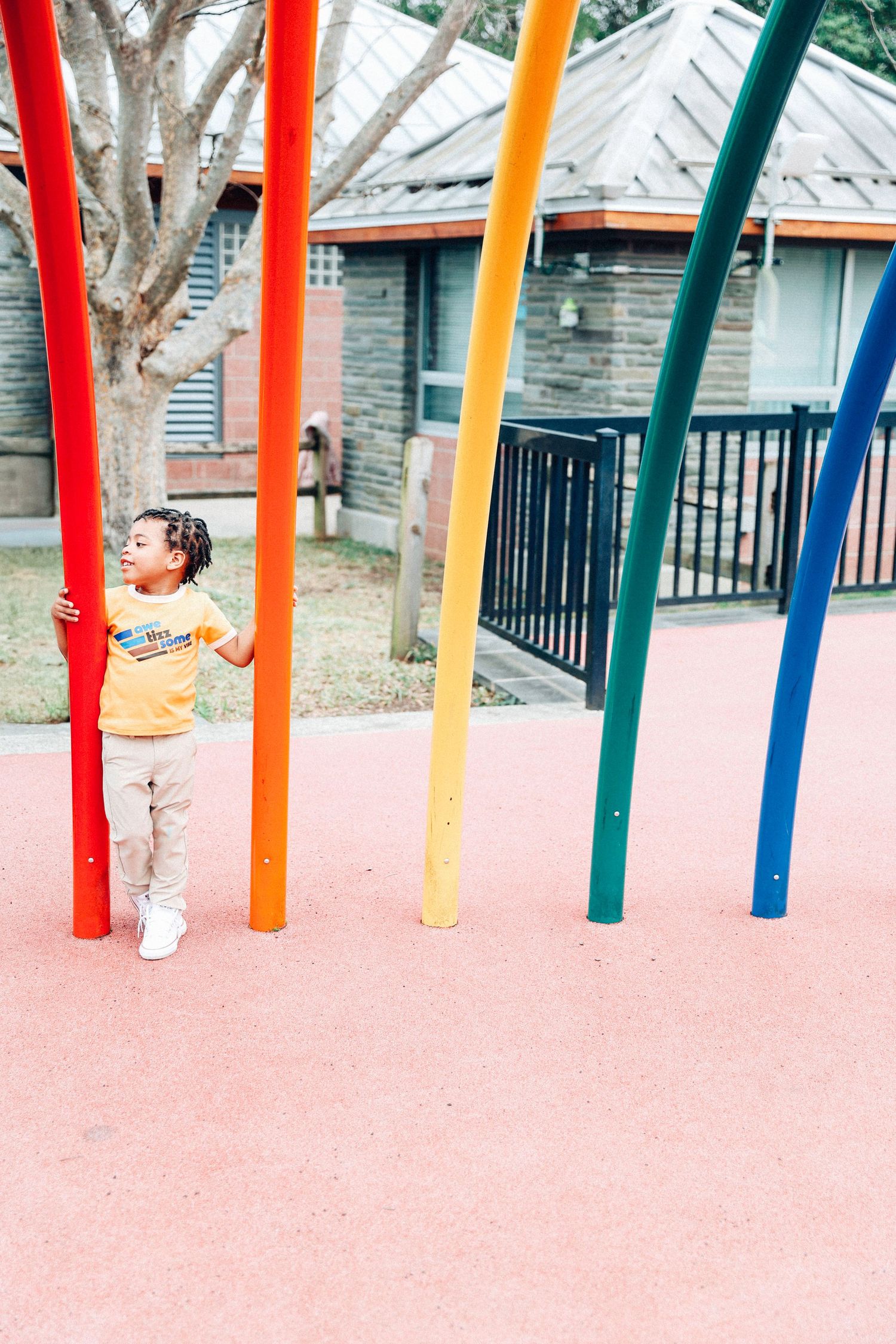 Small child stands among colorful vertical poles on a pink playground surface near a house.