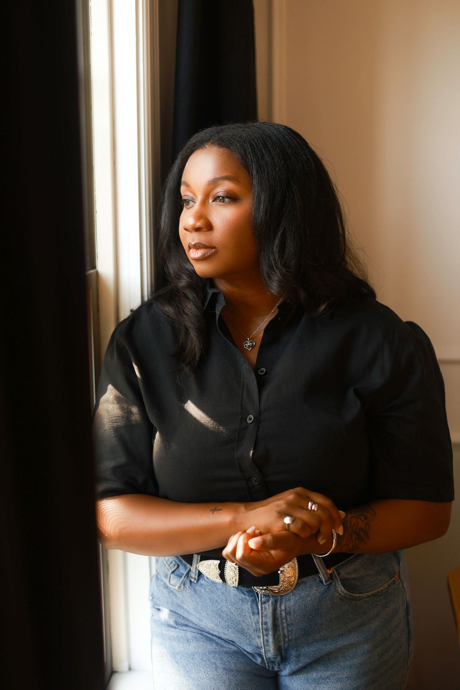 Portrait of someone in a black button-down shirt and jeans looking thoughtfully through a window in natural light.