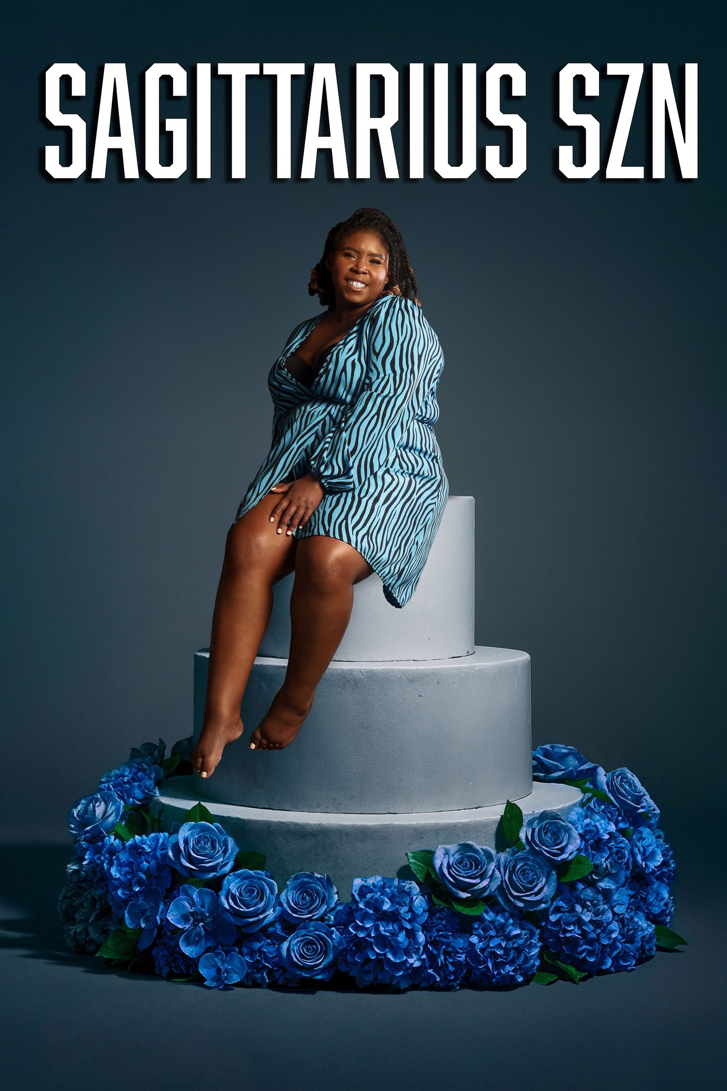 Fashion model poses on white platform surrounded by vibrant blue roses in dramatic studio lighting setup.