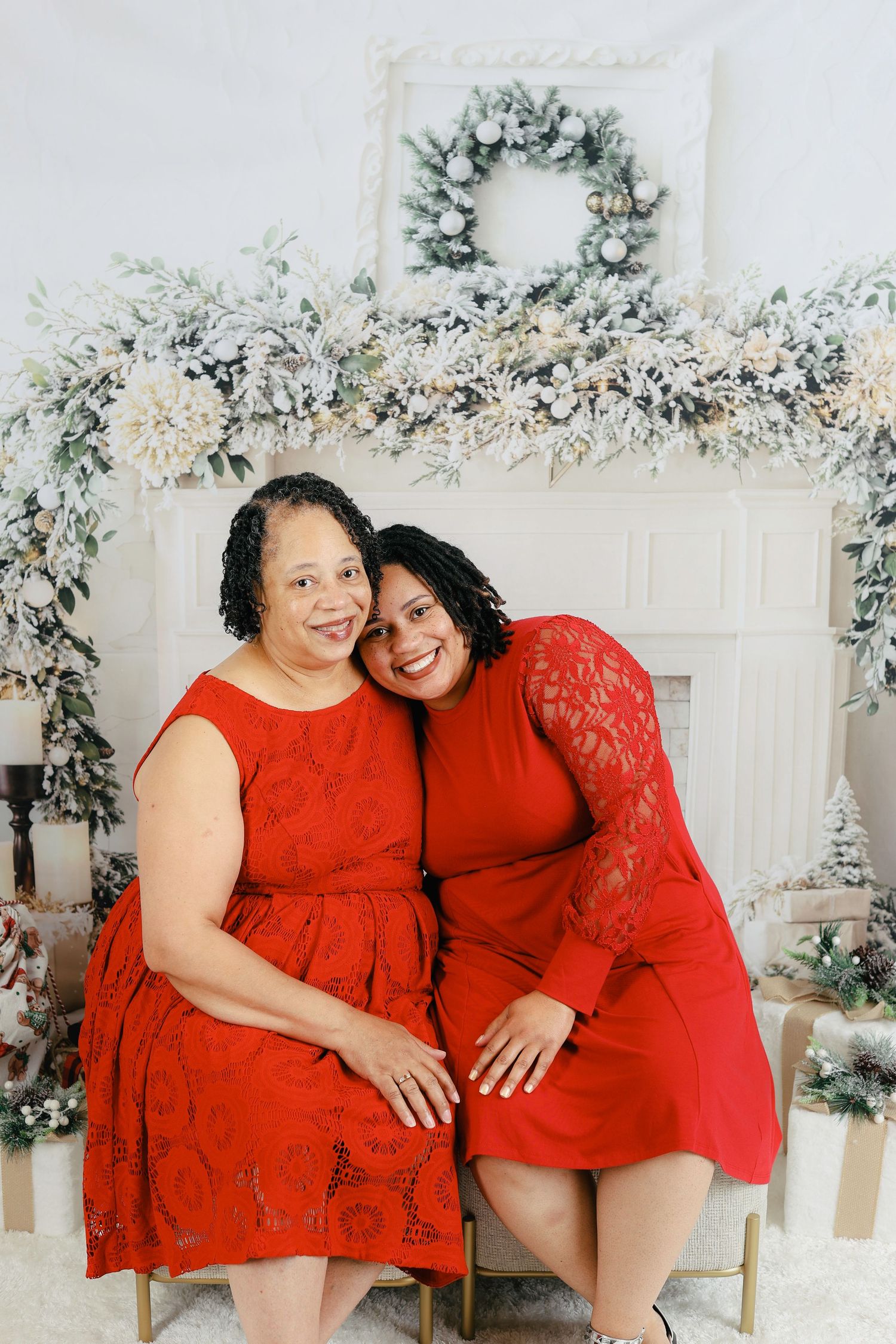 Two people in matching red lace dresses pose together in a winter wonderland studio setting with floral decorations.
