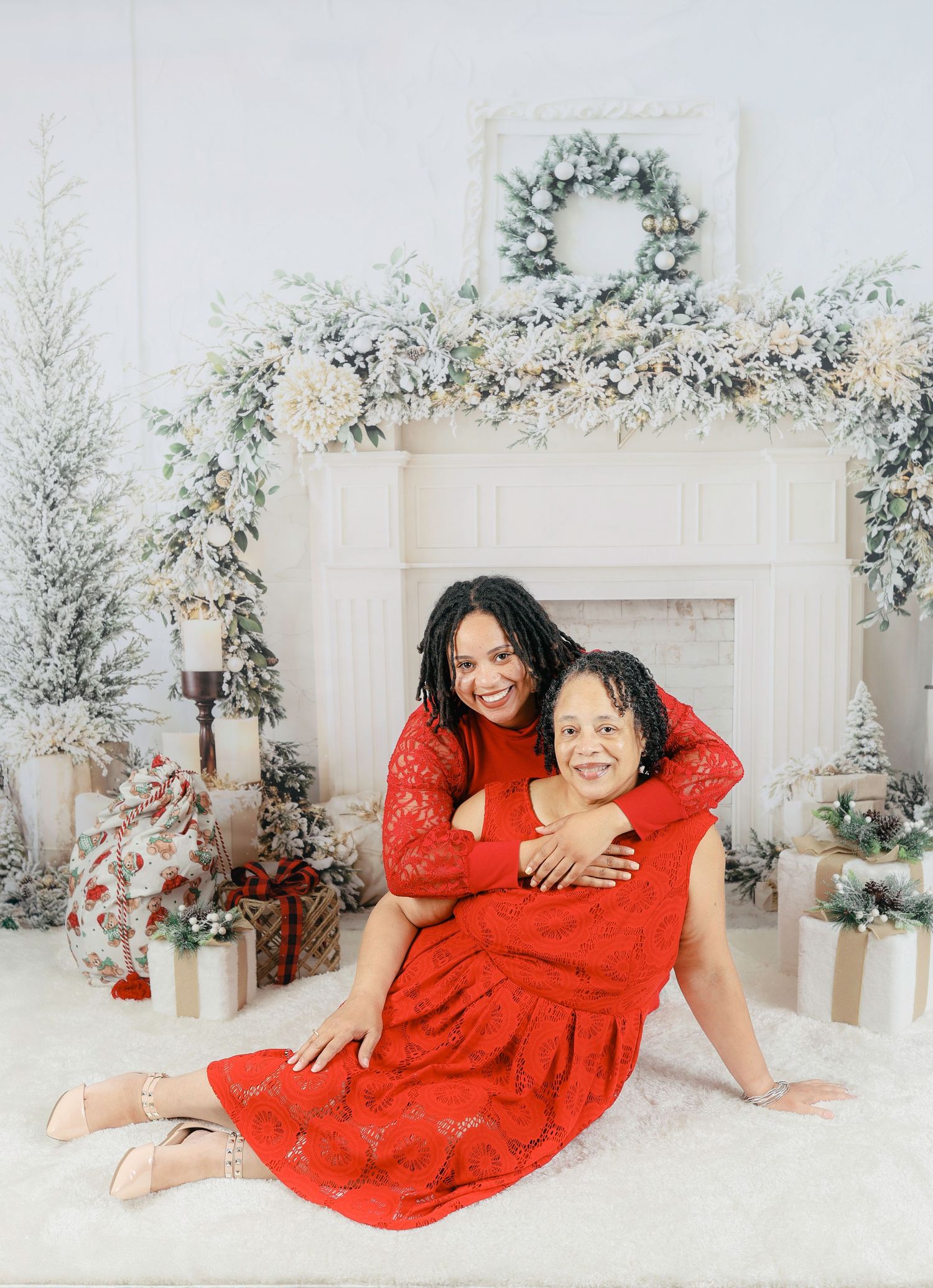 Two people in red dresses strike playful poses on white floor with winter-themed backdrop and decorative flowers.