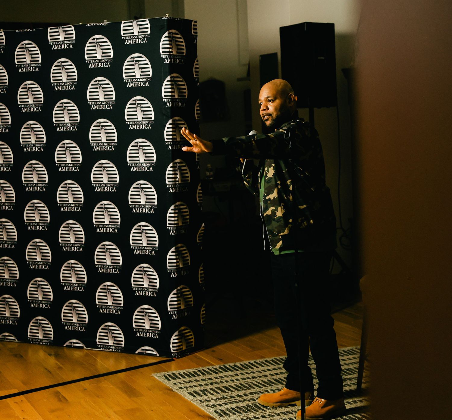 A performer wearing a black jacket and camouflage shirt speaks on stage with a branded repeating pattern backdrop.