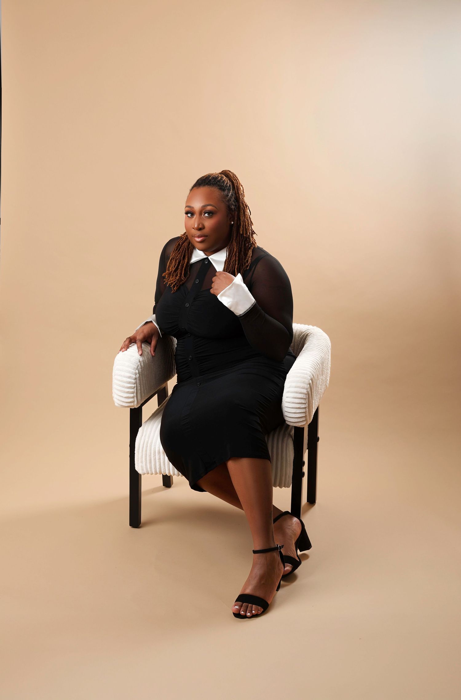 A figure in a black dress and white sleeves poses dramatically on a wooden chair against a beige background.