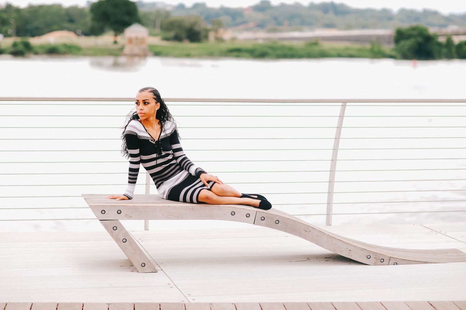 Person in striped outfit posing on a modern waterfront bench with railing and water view.