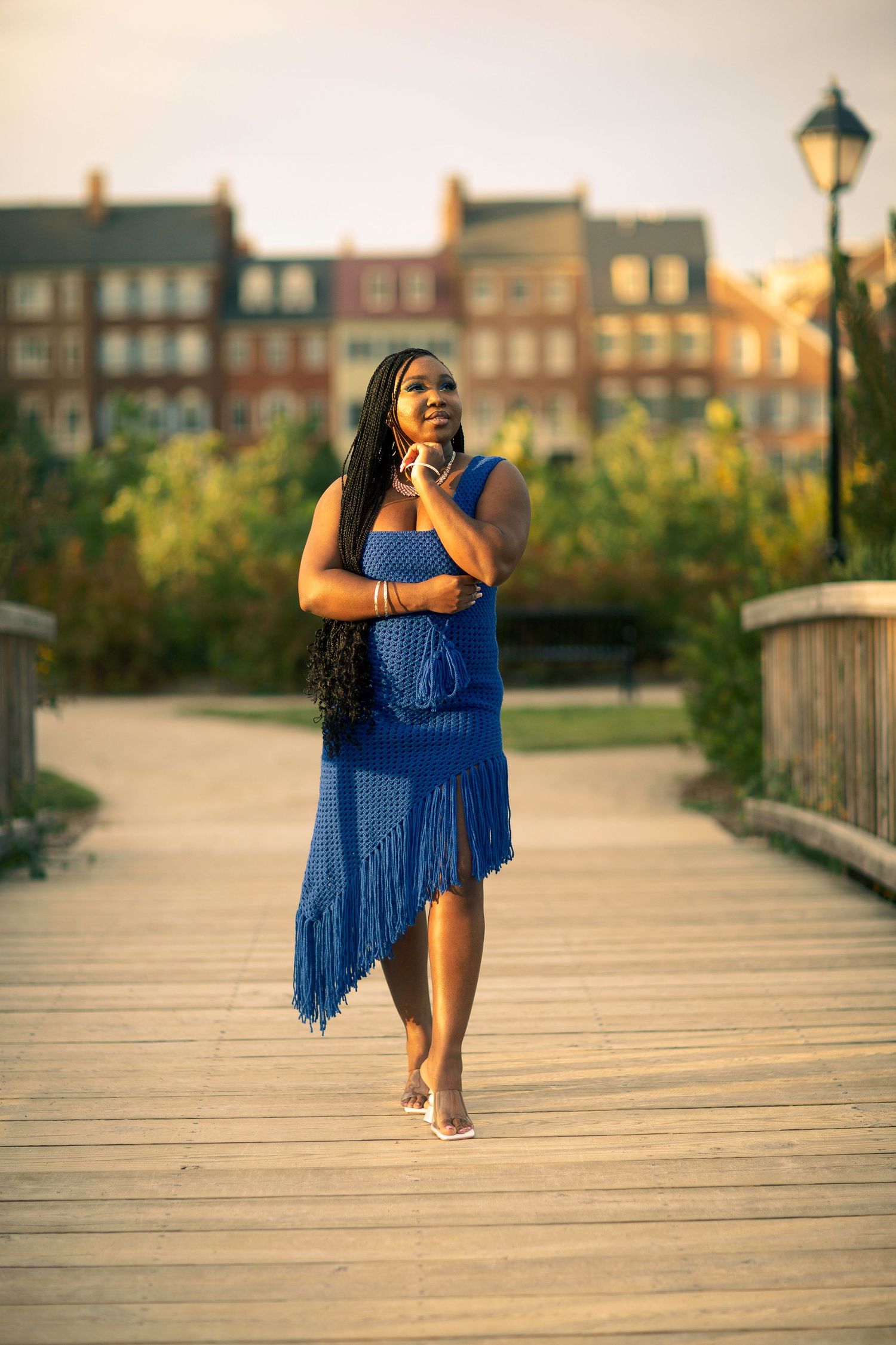 A person in a flowing blue fringe dress stands on a wooden boardwalk at sunset with city buildings in the background.