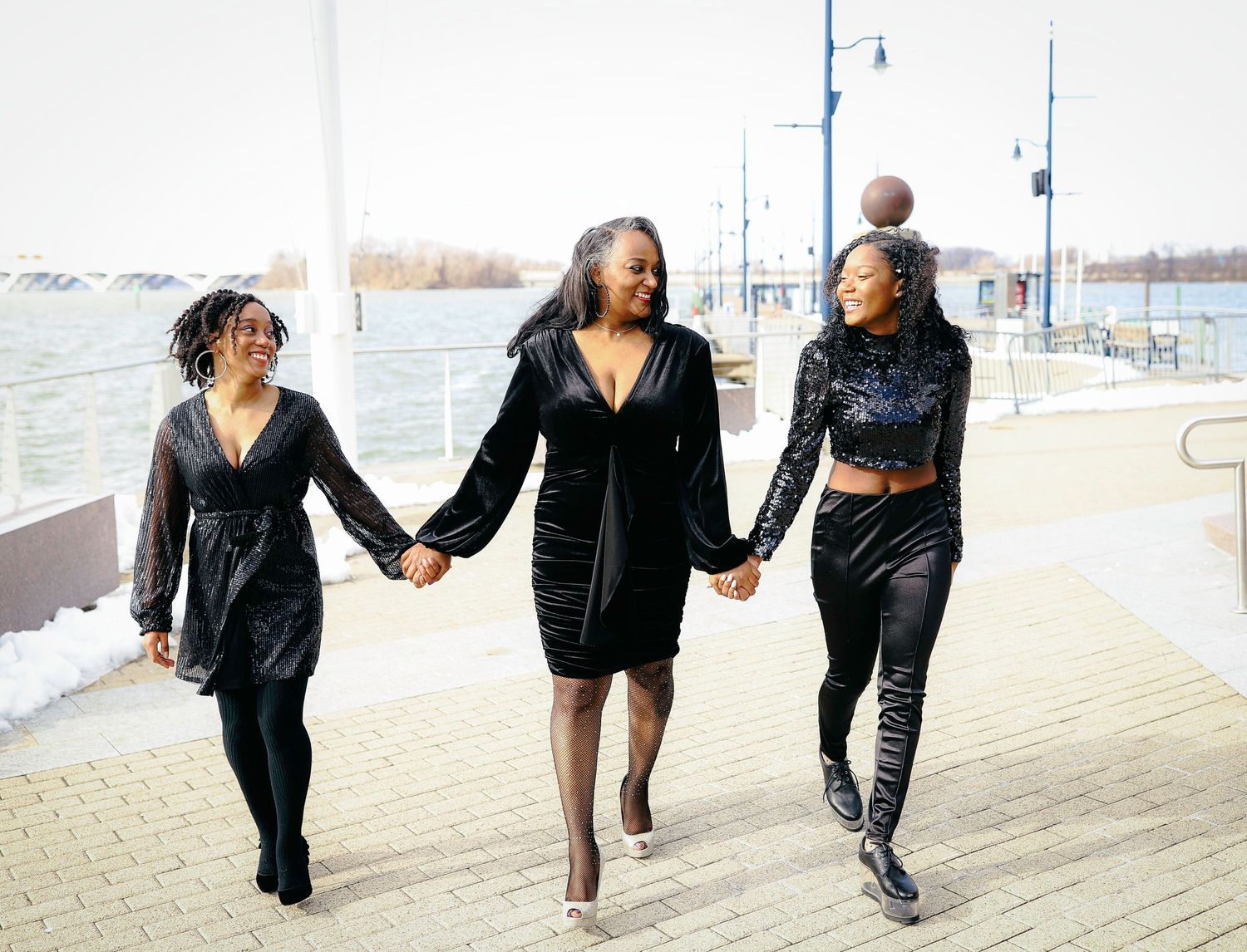 Three friends holding hands while walking and laughing along a waterfront boardwalk.