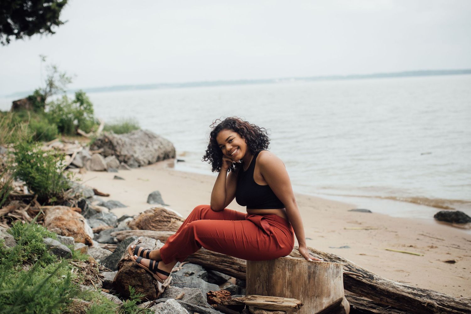 Person in casual attire sitting on driftwood at a rocky beach shoreline.