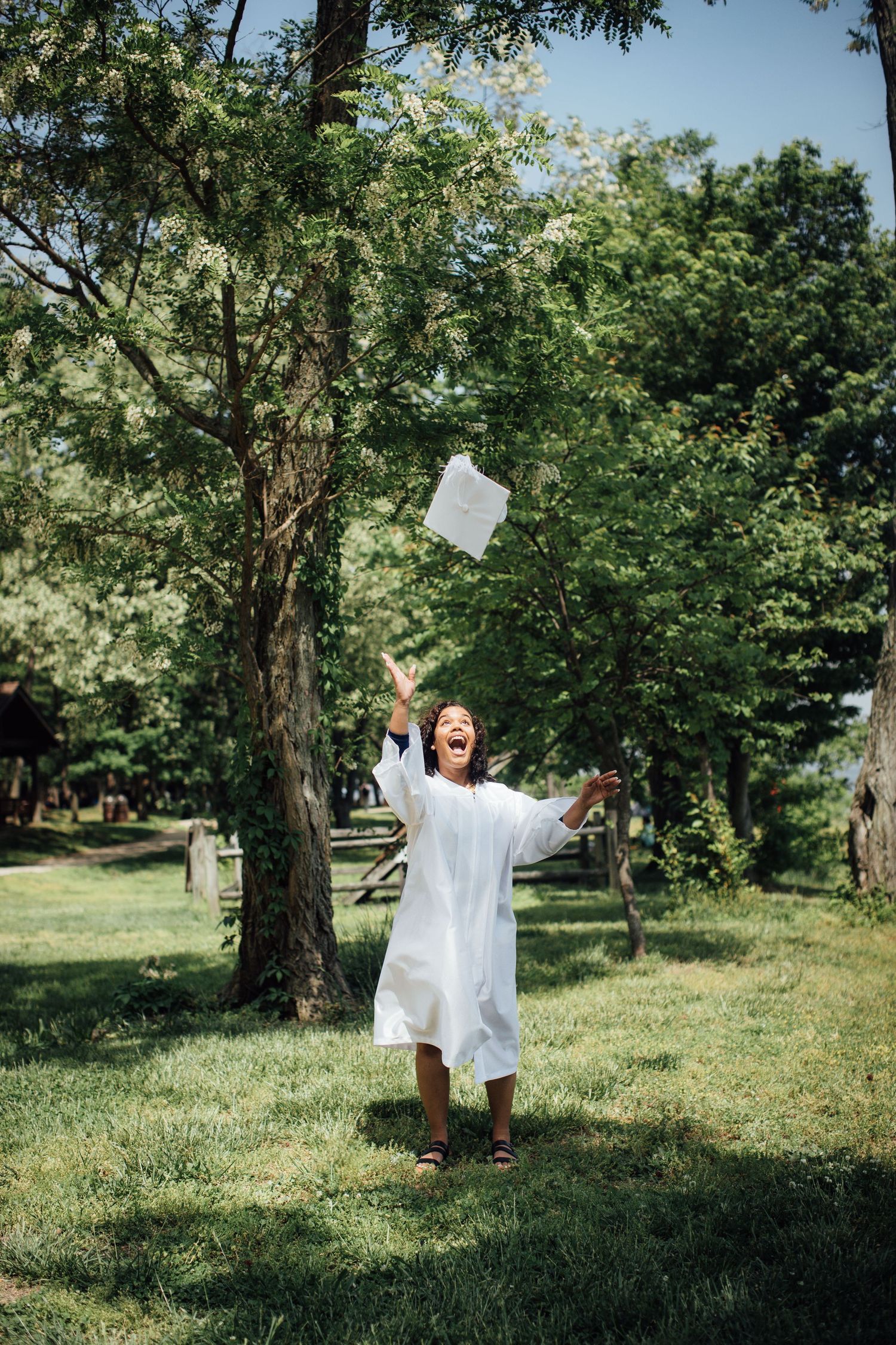 Person in white graduation gown tossing papers in celebration outdoors among trees.