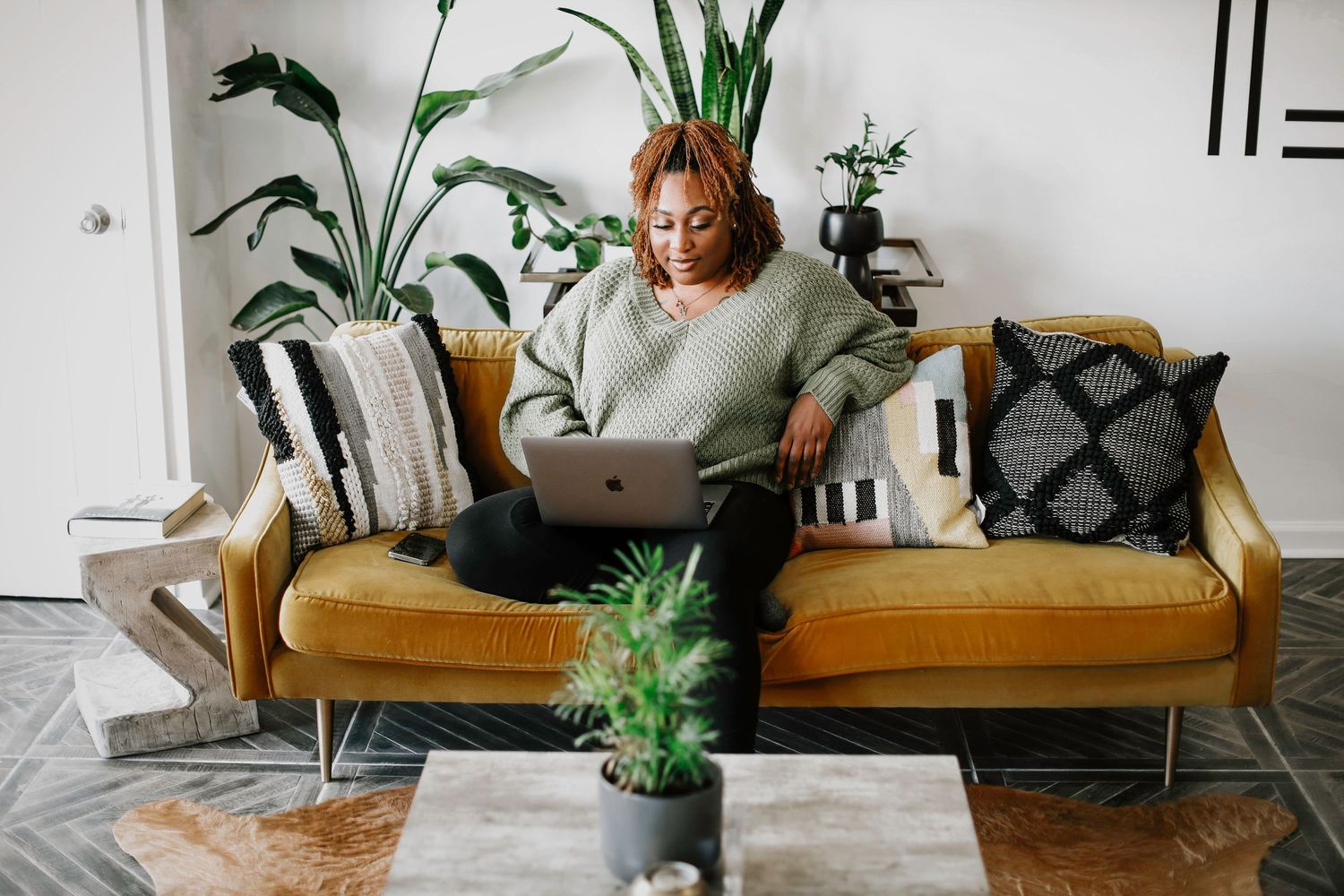 Person working on laptop while sitting on mustard yellow couch with indoor plants.