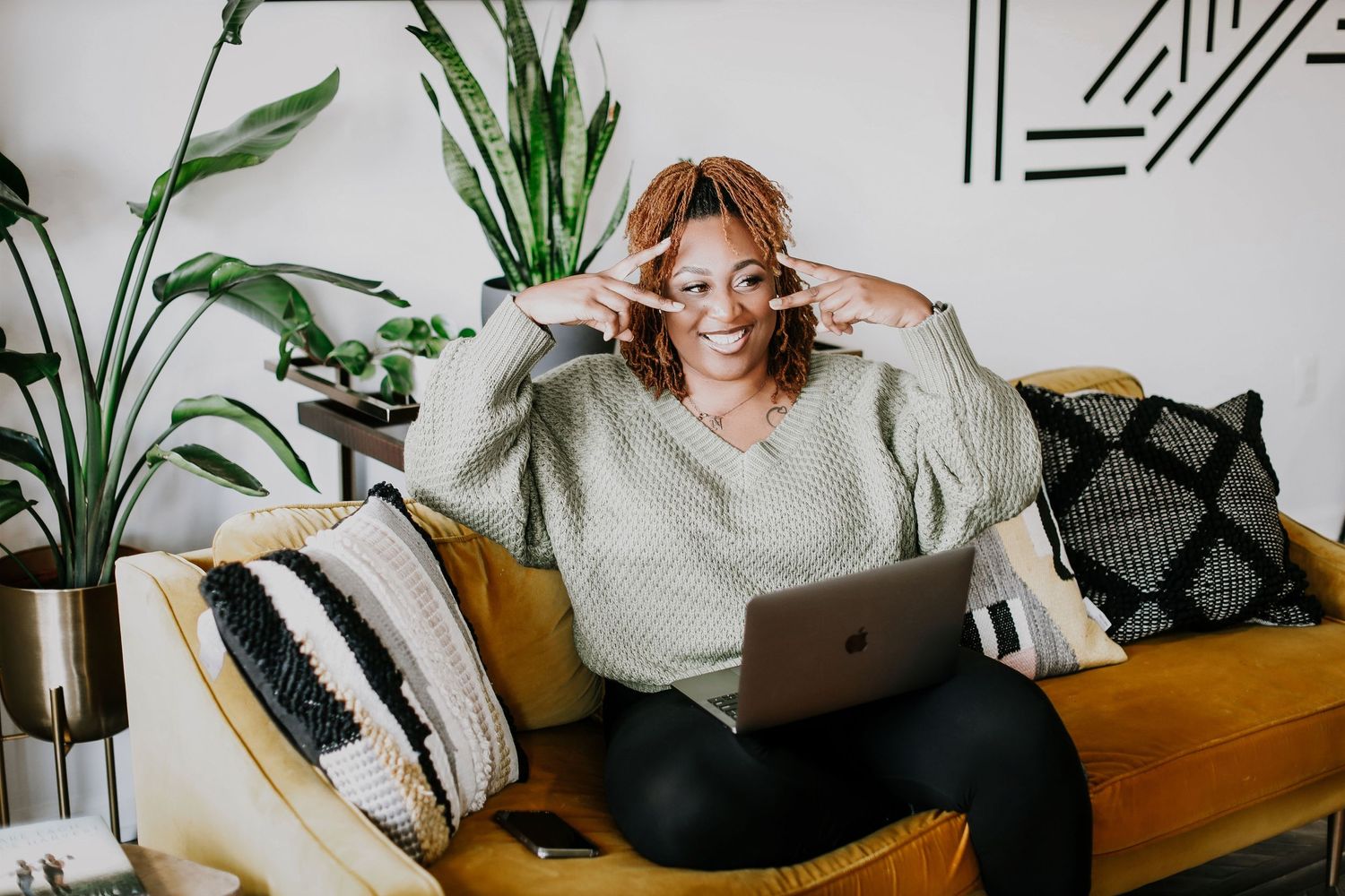 Person in grey sweater adjusting glasses while using laptop on yellow couch with plants.