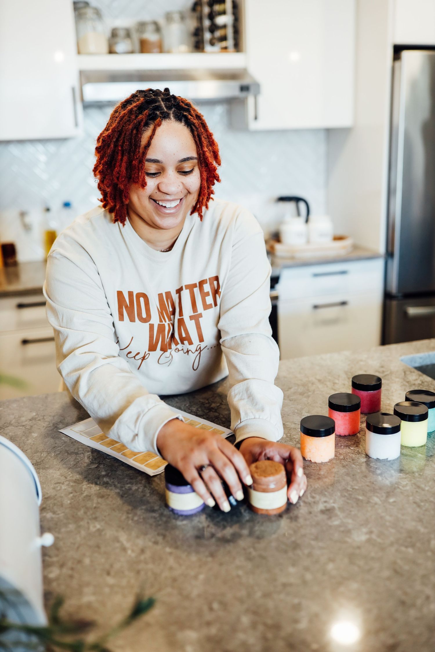 A person in a cream sweatshirt arranges small product containers on a kitchen counter.