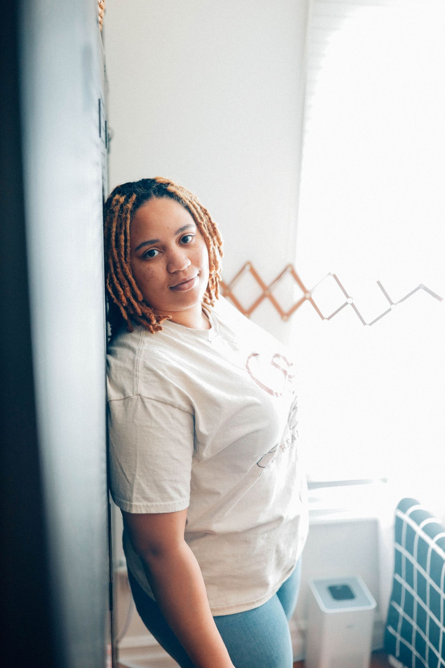 A person with blonde braids leans against a white wall near a window in casual attire.