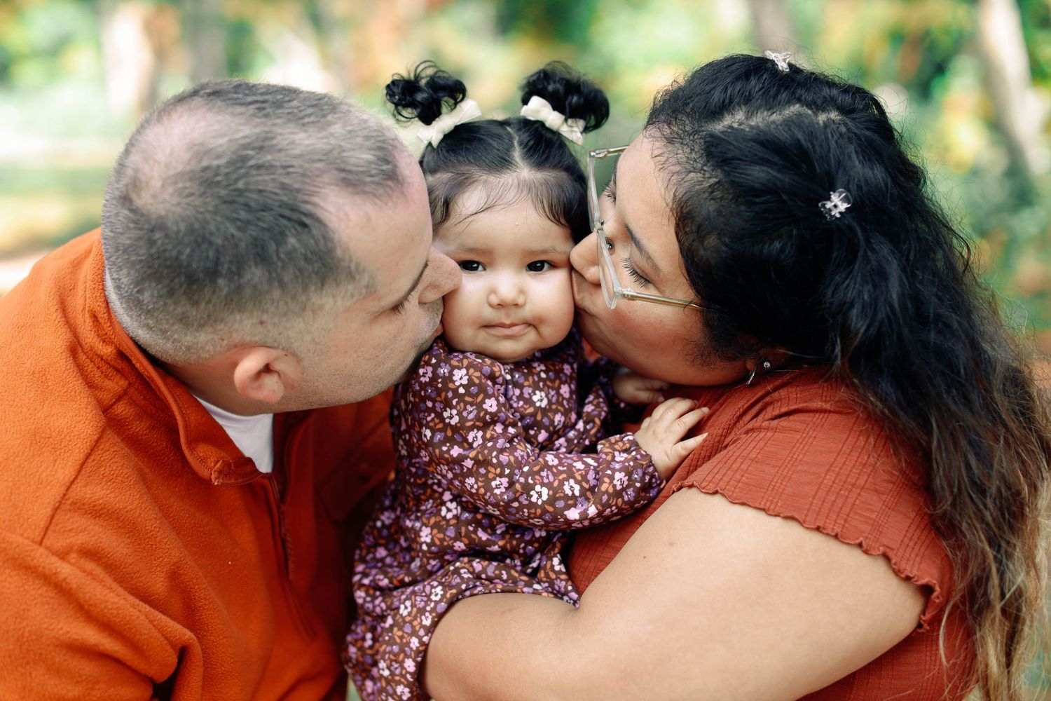 A family moment captured outdoors with parents kissing their baby's cheeks.