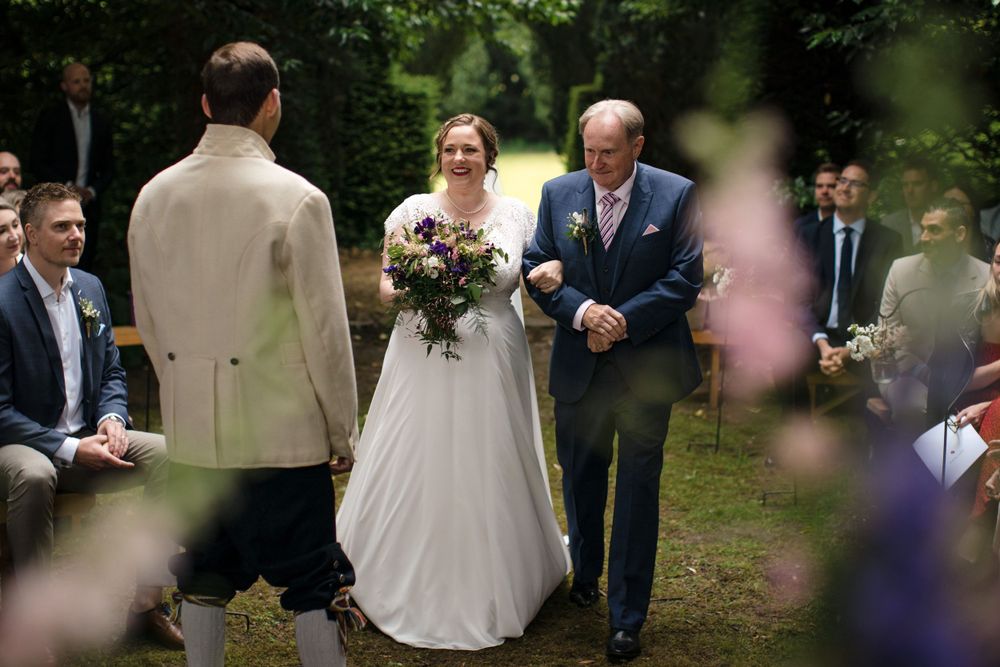 A wedding ceremony takes place at the Tangled Garden in a lush garden setting with attendees gathered around during the processional.