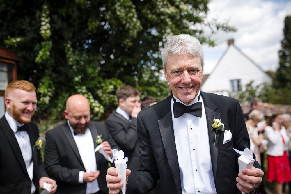 A person in a black tuxedo and bowtie holds a drink while smiling at an outdoor wedding reception at Tangled Garden wedding venue.