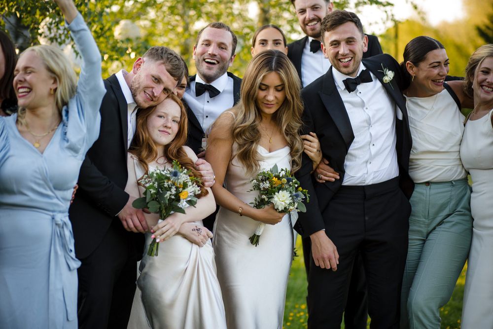 A joyful wedding party in formal attire stands together outdoors laughing and smiling in golden hour light at the Tangled Garden, Grand Pre