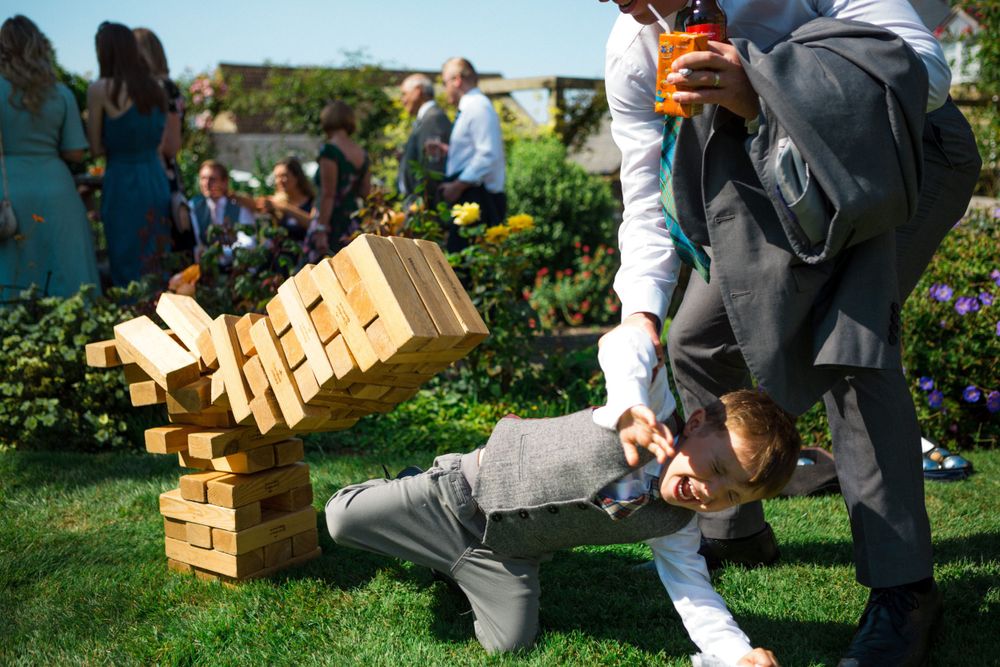 Giant wooden Jenga blocks tumble down on a sunny lawn as people gather during an outdoor wedding at Tangled Garden, Grand Pree venue