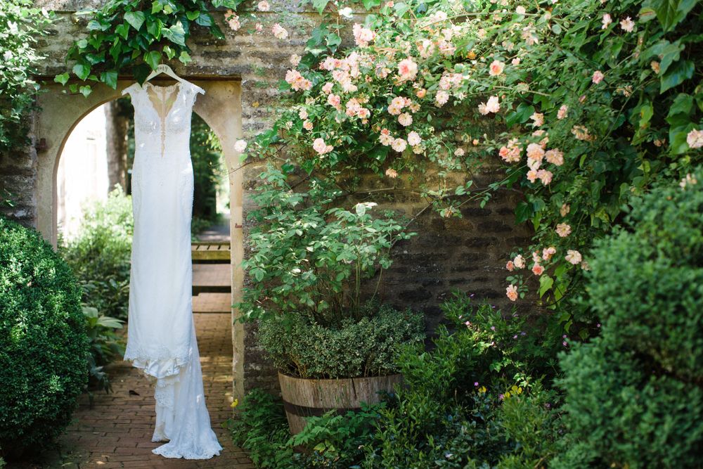 An elegant white wedding dress hangs in a garden archway at the Tangled Garden surrounded by climbing roses and greenery.