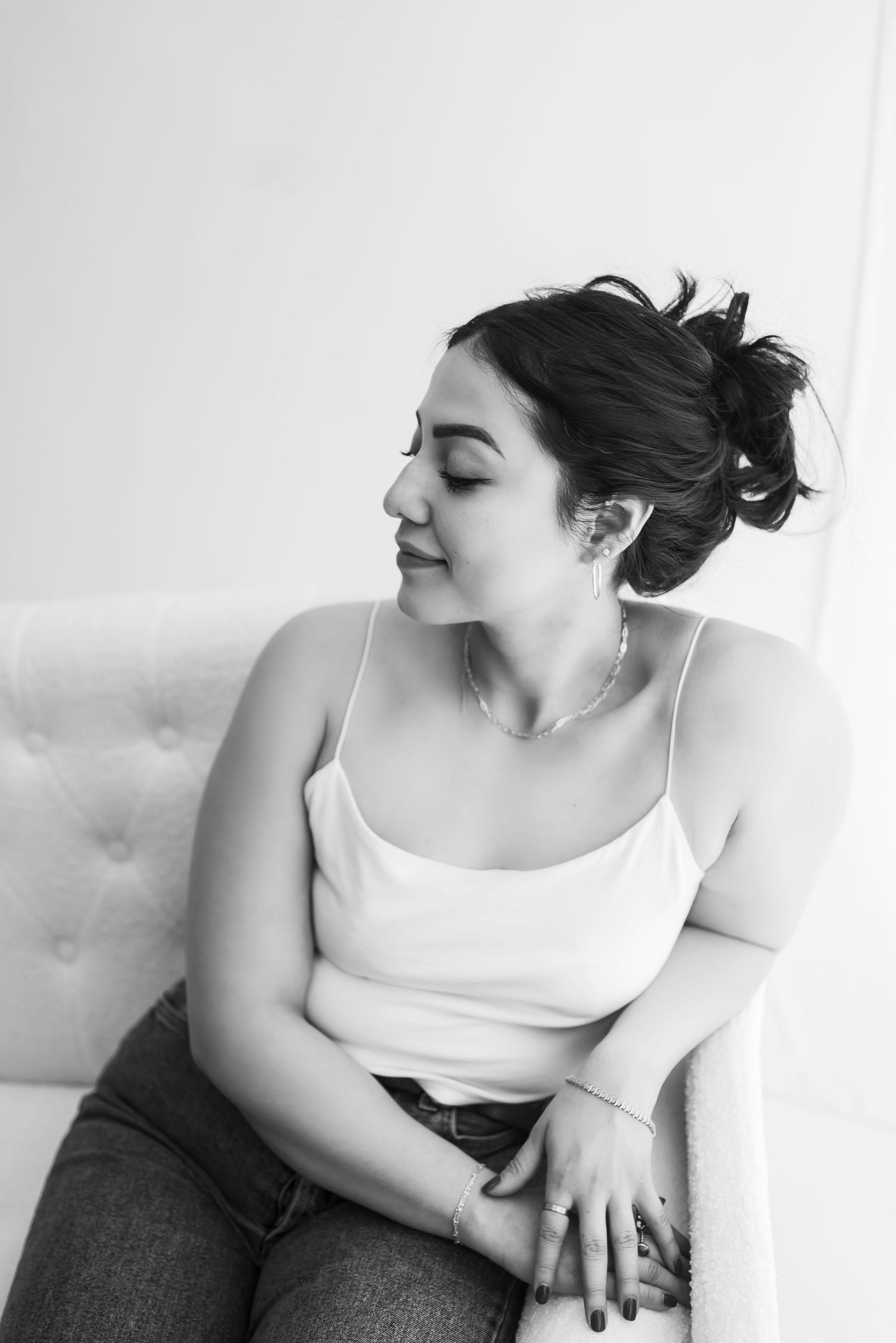 Black and white portrait of woman in white tank top and messy bun sitting on couch.