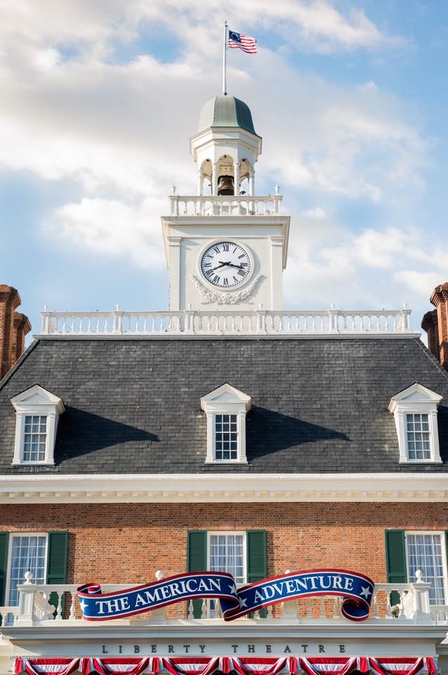 A colonial-style building with a clock tower cupola, American flag, and 'The American Adventure' sign on brick facade.