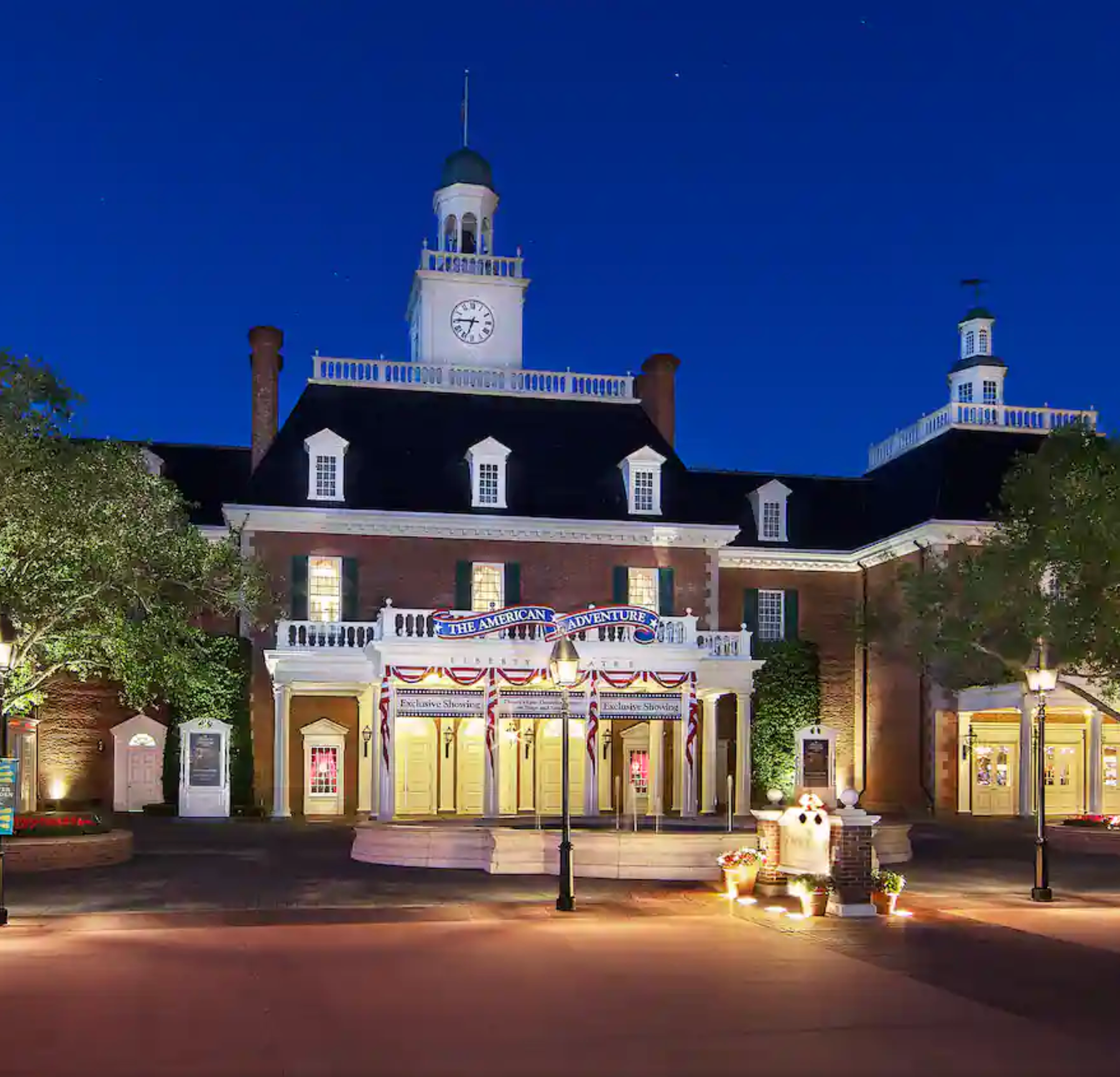 Historic Colonial Williamsburg buildings illuminated at night with warm lighting against a deep blue evening sky.