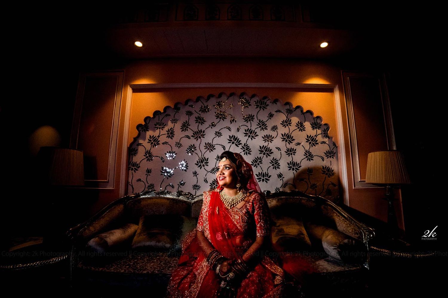 A dramatic bridal portrait in front of an ornate wall with someone wearing a luxurious red traditional dress in low lighting.