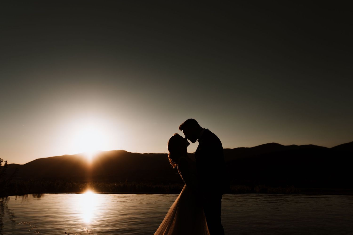 A silhouetted couple embraces against a stunning sunset over a lake with mountains in the background.