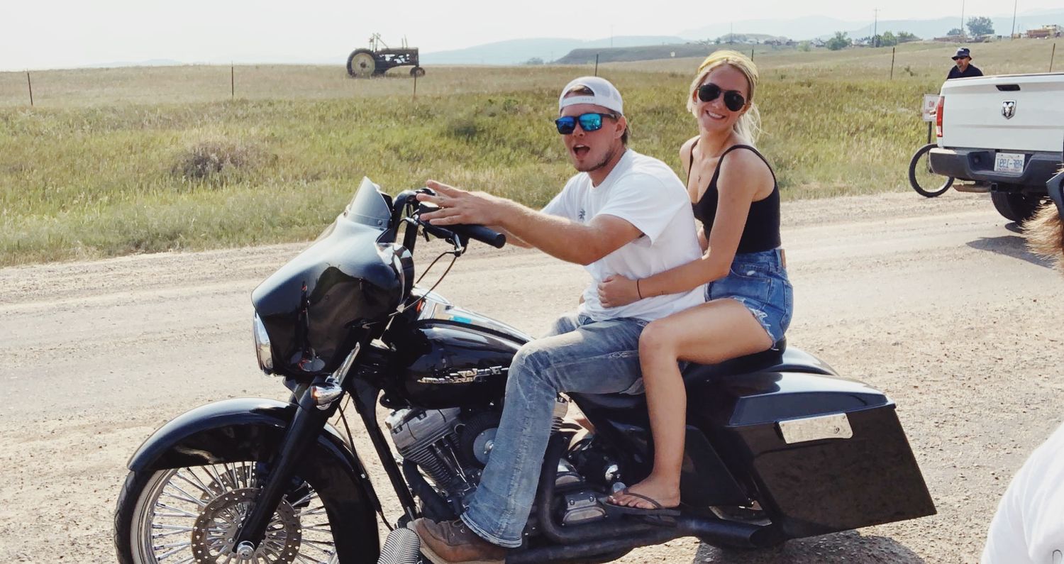 Two people riding a motorcycle on a dirt road through open grassland on a sunny day.