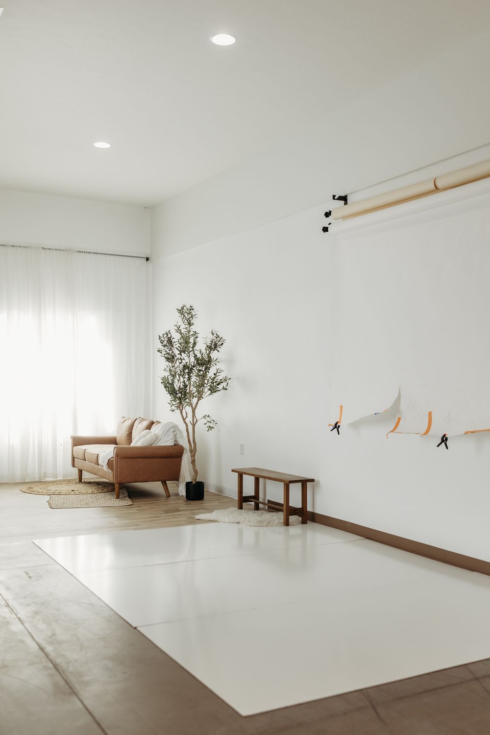 Minimalist photography studio with white walls, leather sofa, olive tree, wooden bench, and backdrop rail system.