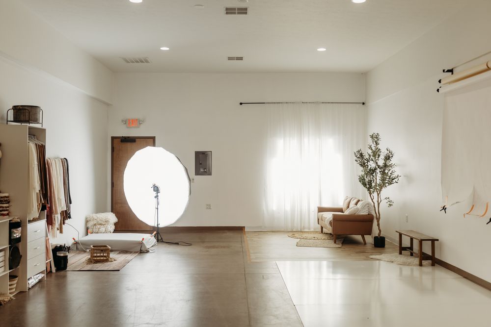 Bright minimalist photography studio with ring light, sheer curtains, potted tree, and neutral decor.