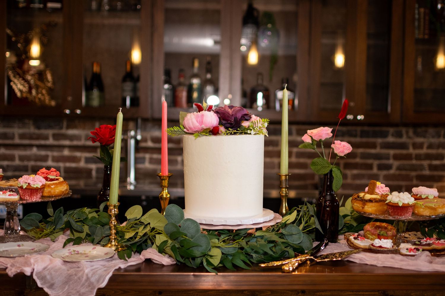 White wedding cake surrounded by candlesticks and fresh flowers on rustic wooden table with dessert display.