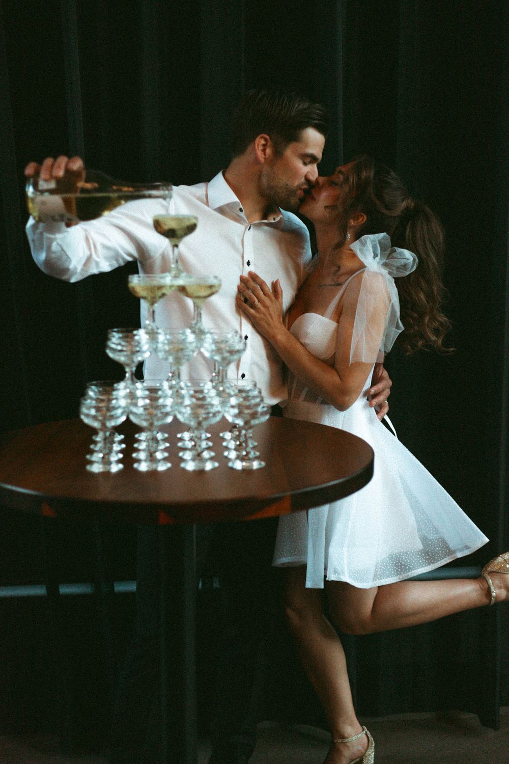 Romantic couple pouring champagne into tower of glasses while embracing near dark wooden table.