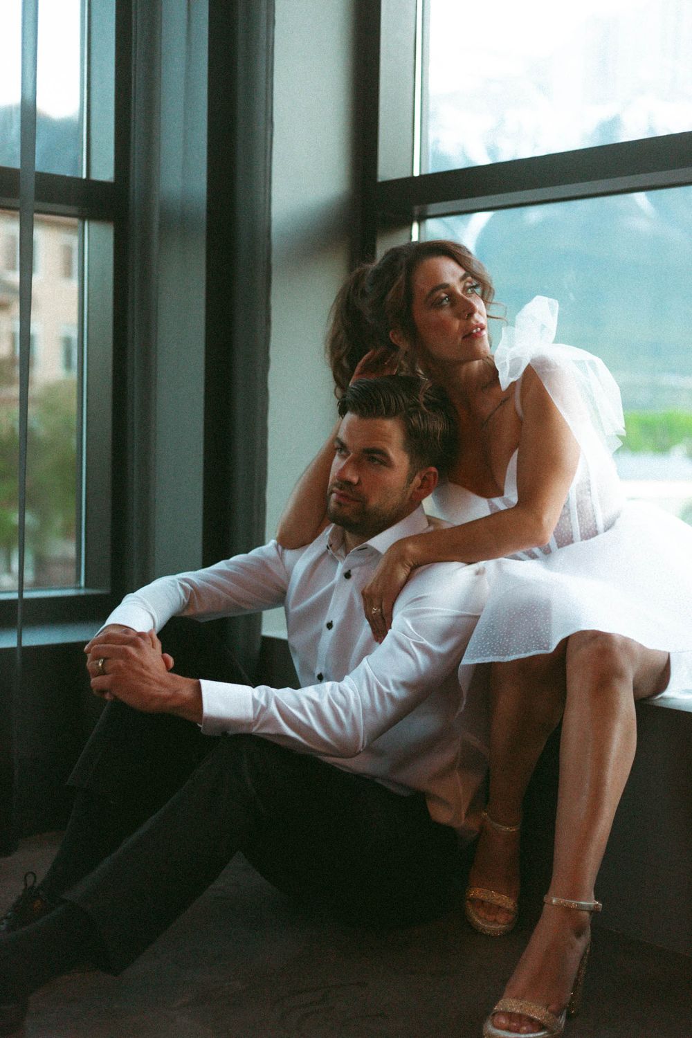 A romantic couple in wedding attire pose intimately near large windows with natural light streaming in.