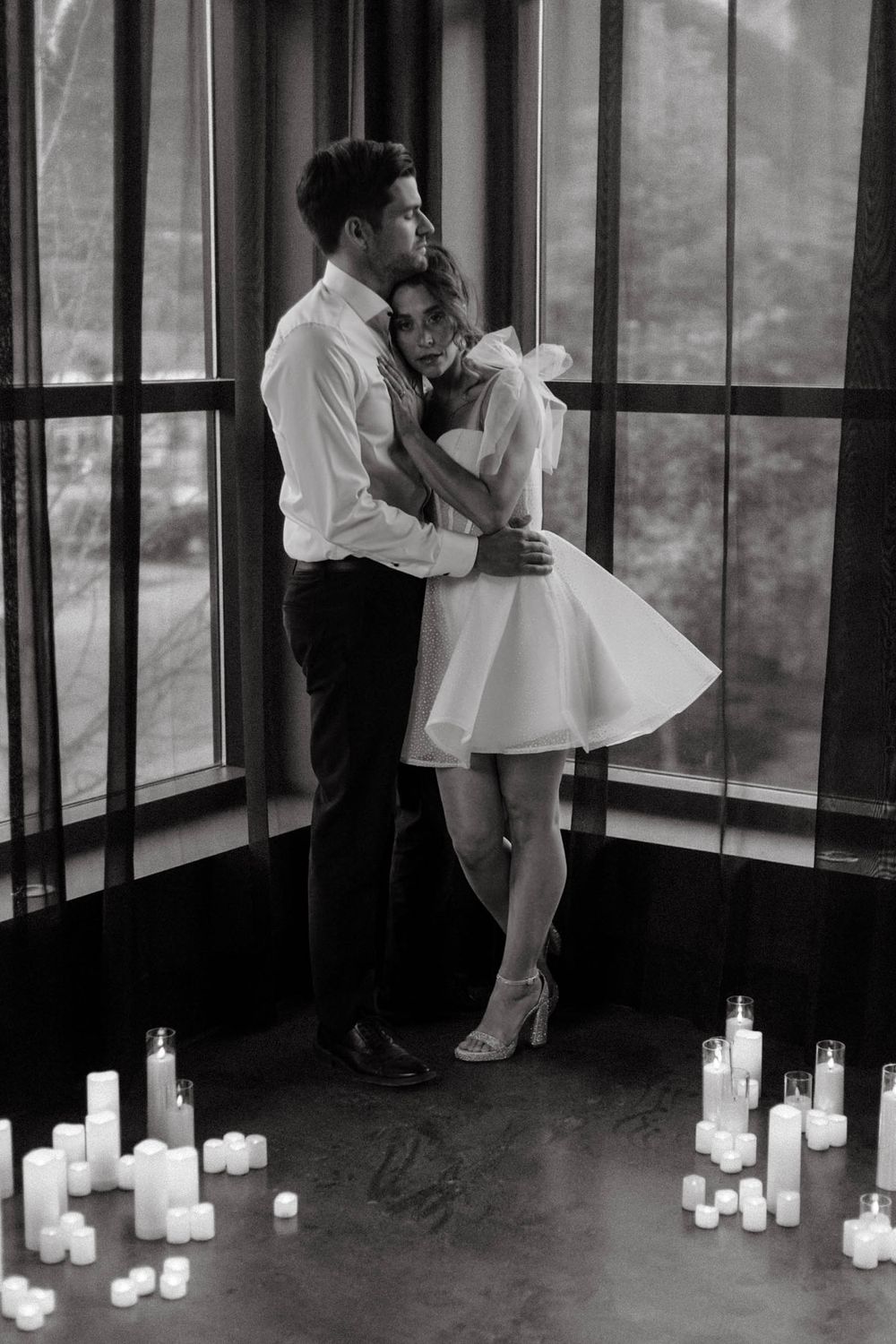 A romantic black and white photo of a couple embracing by a window with glowing candles scattered on the floor.