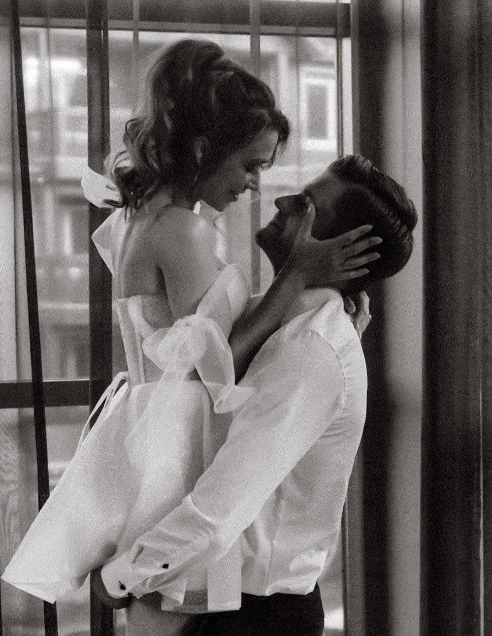 A romantic black and white photograph shows a couple in an intimate embrace by a window, dressed in white clothing.