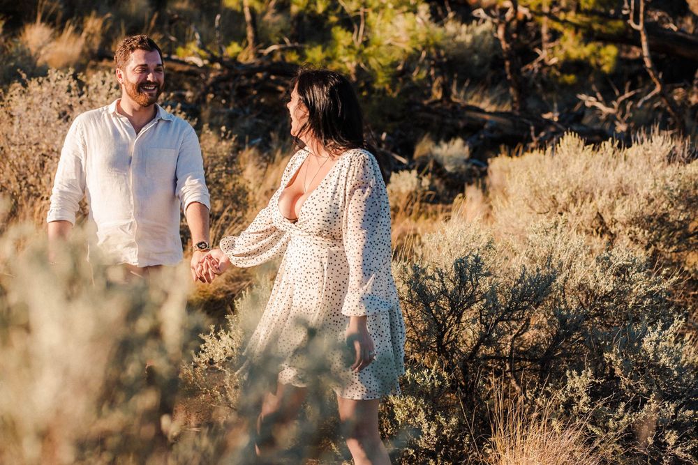 A couple shares a romantic moment while walking through tall grass at golden hour in a desert landscape.