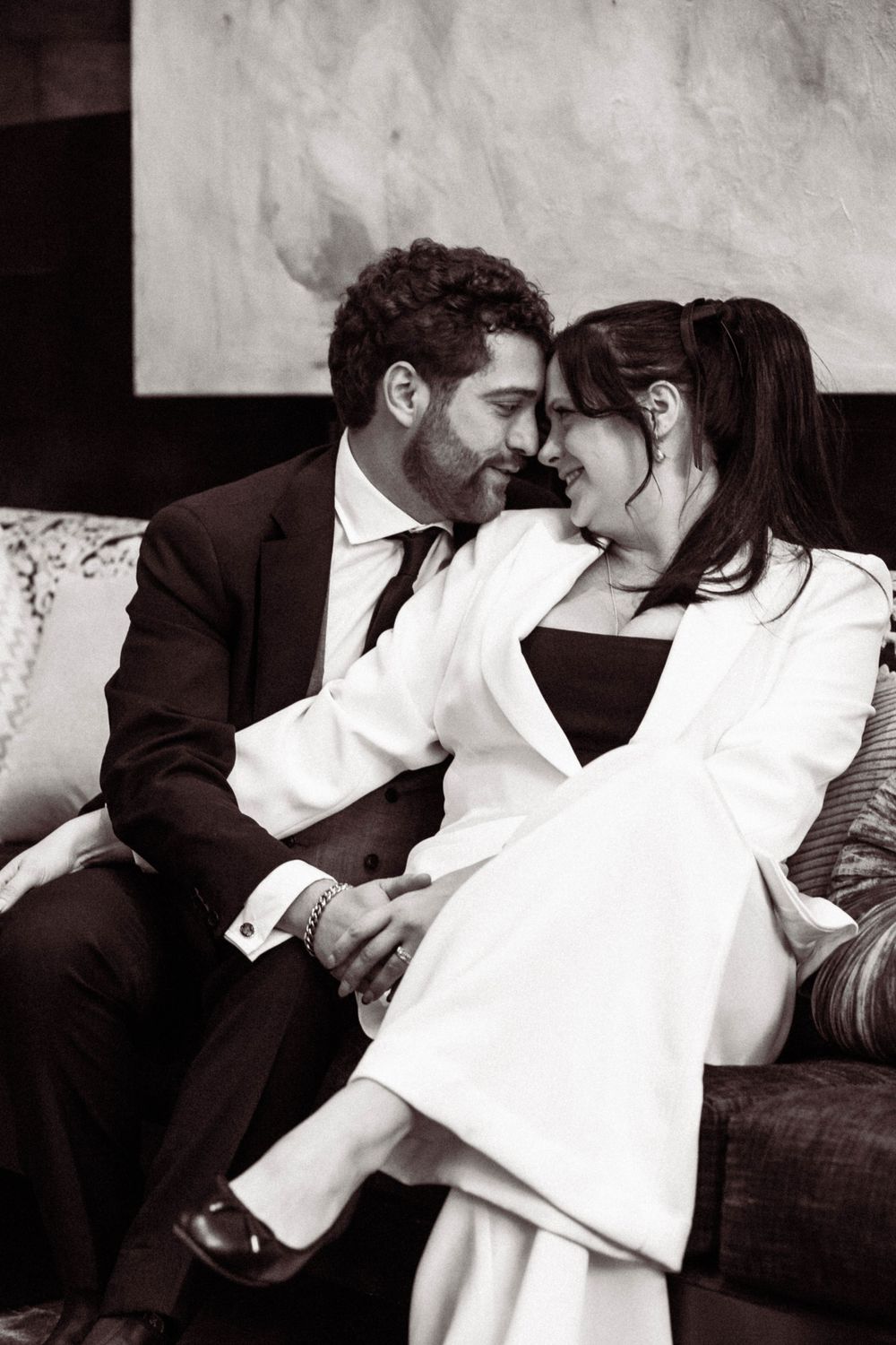 An intimate black and white portrait of a couple in formal attire sharing a tender moment on a couch.