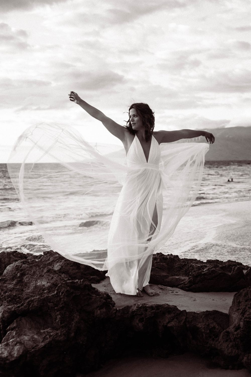 A figure in a flowing white dress stands gracefully on coastal rocks as ocean waves crash in the background at sunset.