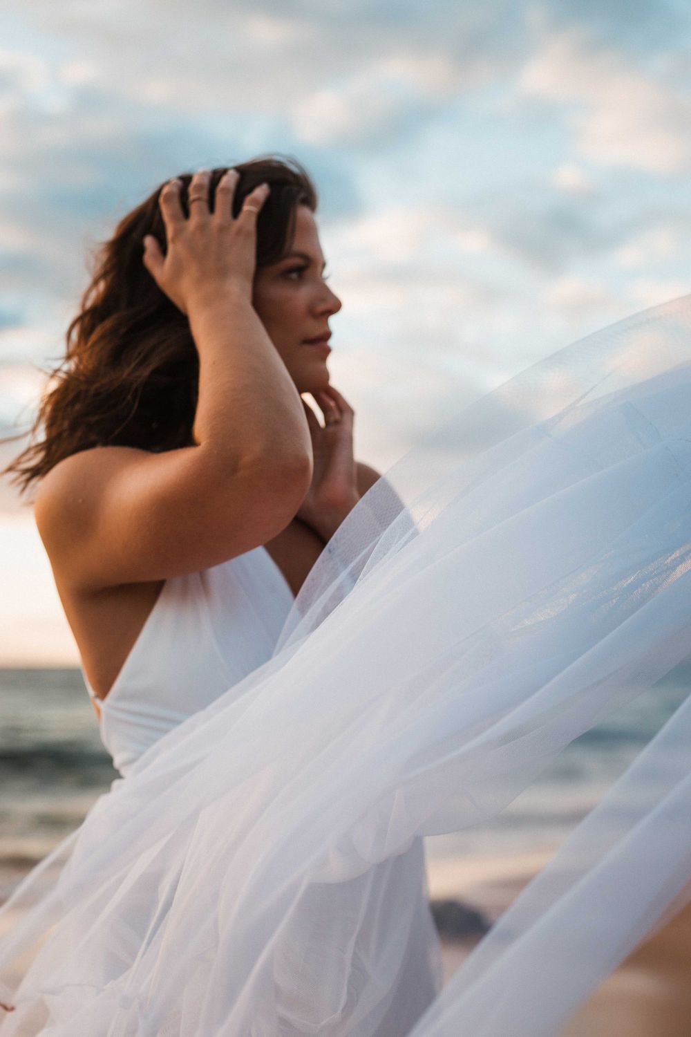A person in a flowing white dress stands on a beach at sunset with wind blowing through their hair.