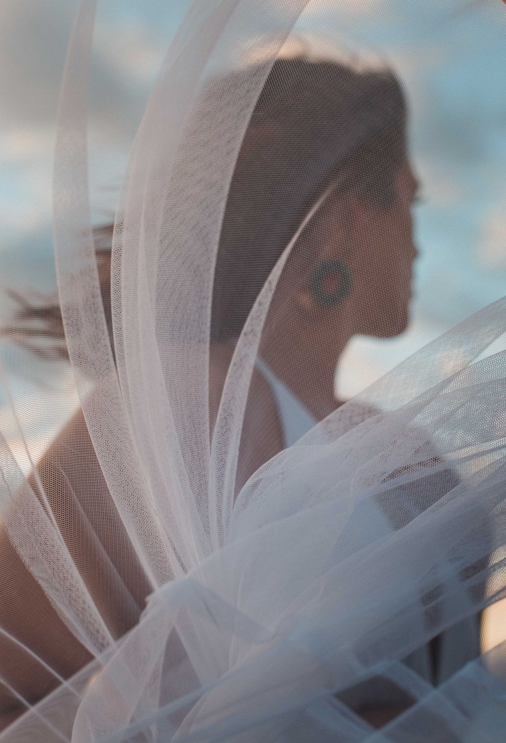 A delicate white wedding veil catches the sunlight as it flows in the breeze against a soft blue sky background.