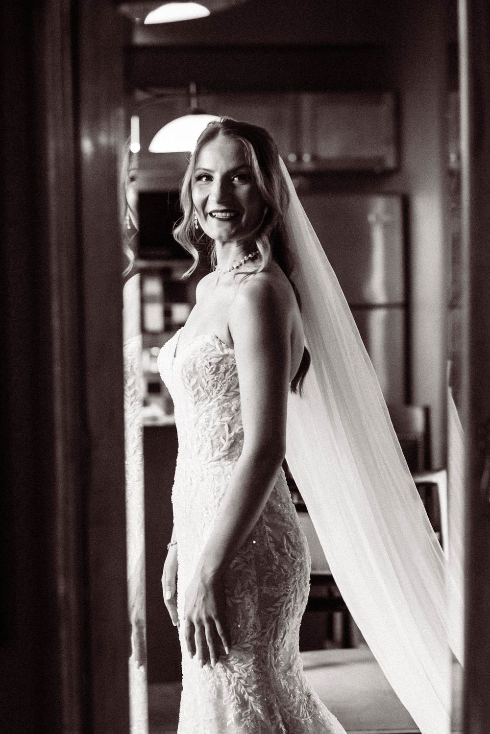 Black and white portrait of a bride in a fitted lace wedding gown with flowing veil in elegant indoor setting.