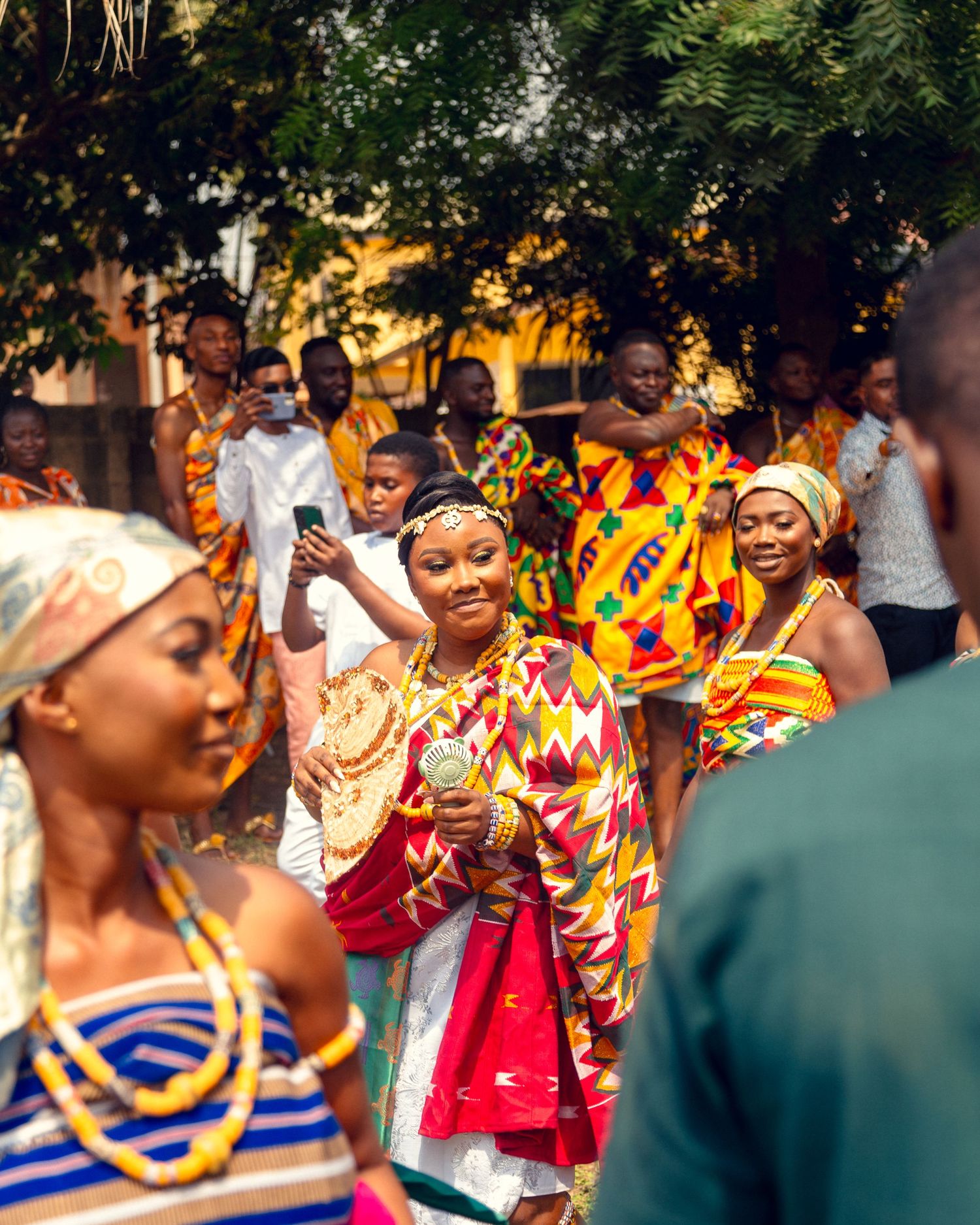 People in colorful traditional Ghanaian kente cloth gather at an outdoor celebration during sunset.