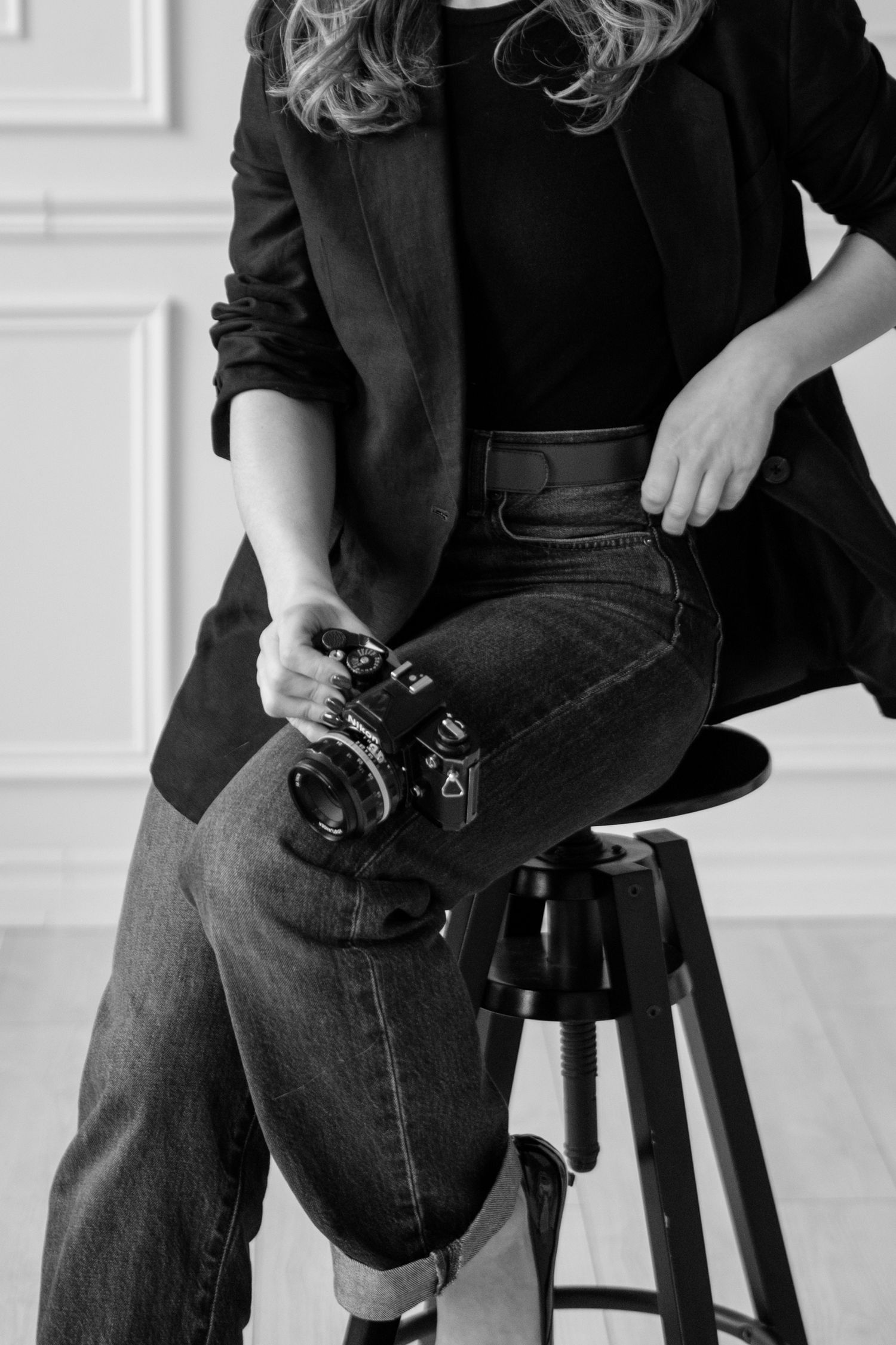 A person in dark clothing sits on a stool holding a vintage camera in a minimalist white studio setting.
