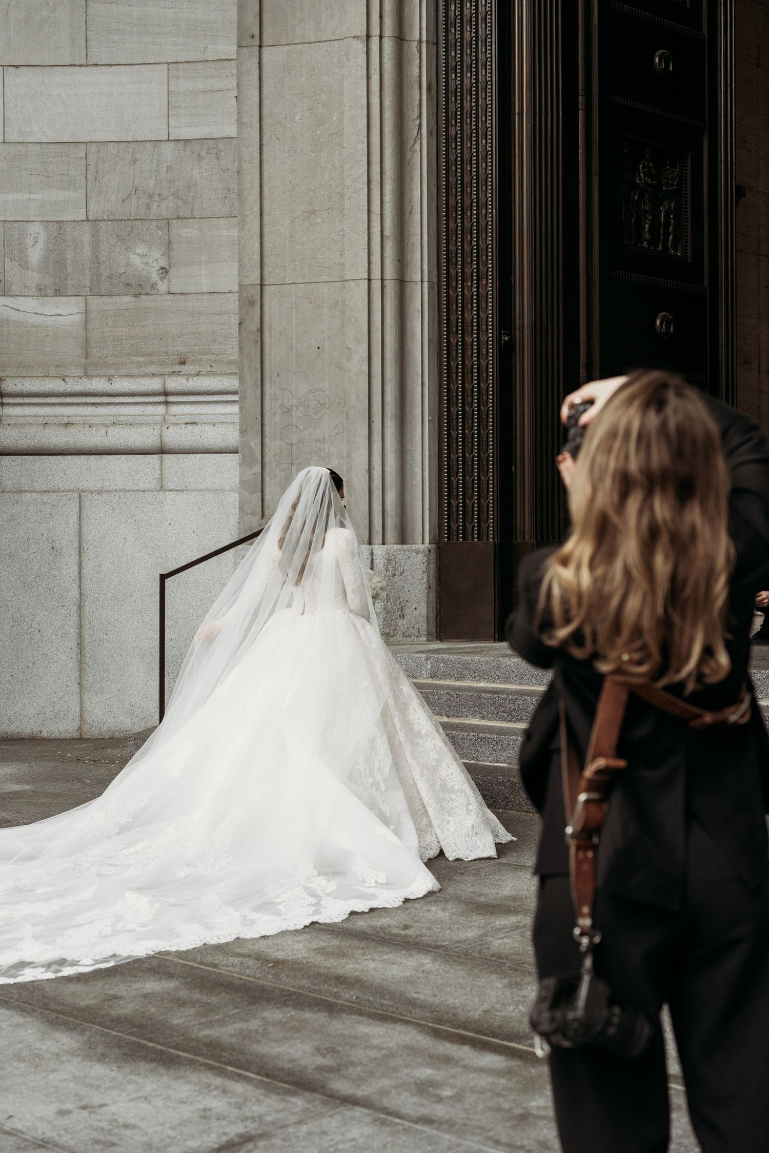 Behind-the-scenes shot of a flowing white wedding dress train entering a grand stone building.
