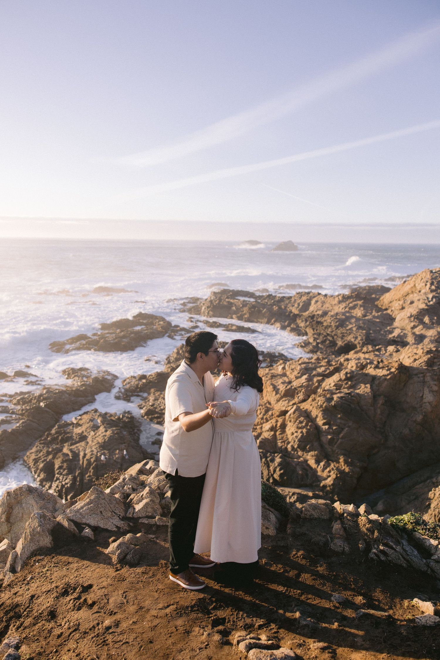 A couple stands together on dramatic rocky cliffs overlooking the misty Pacific Ocean coastline at sunset.