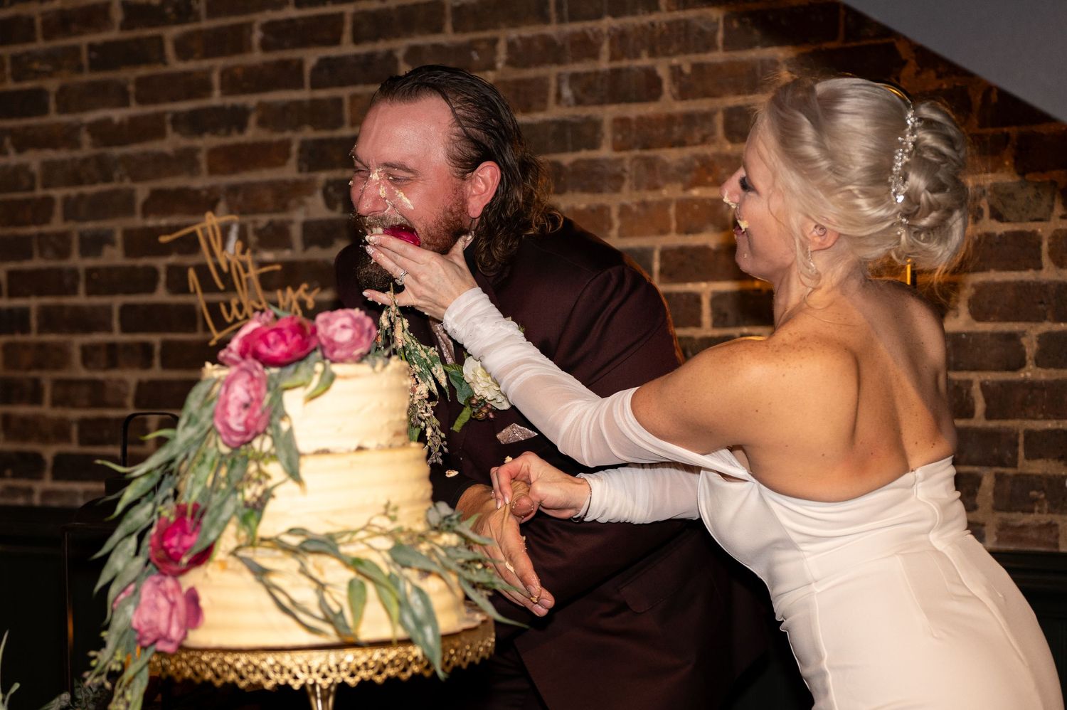 A happy couple shares a playful moment while cutting their floral-decorated wedding cake in a brick-walled venue.