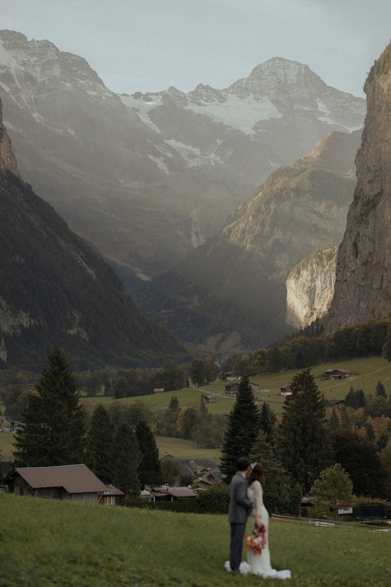 Wedding couple standing in meadow with dramatic Alpine mountains in background.