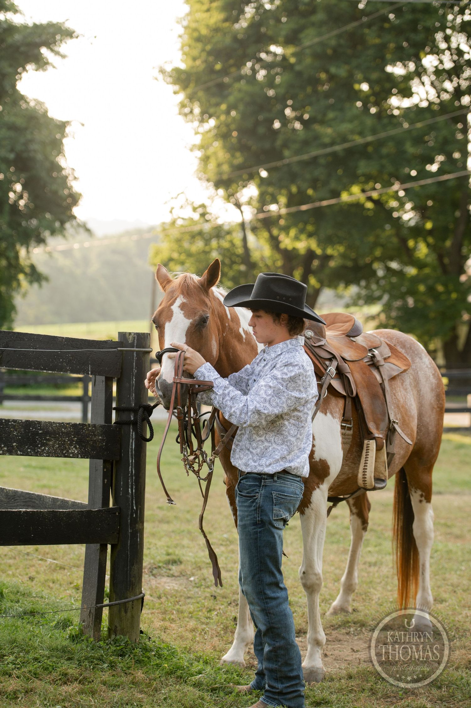 A person in western attire handles a paint horse near wooden fencing at sunset.