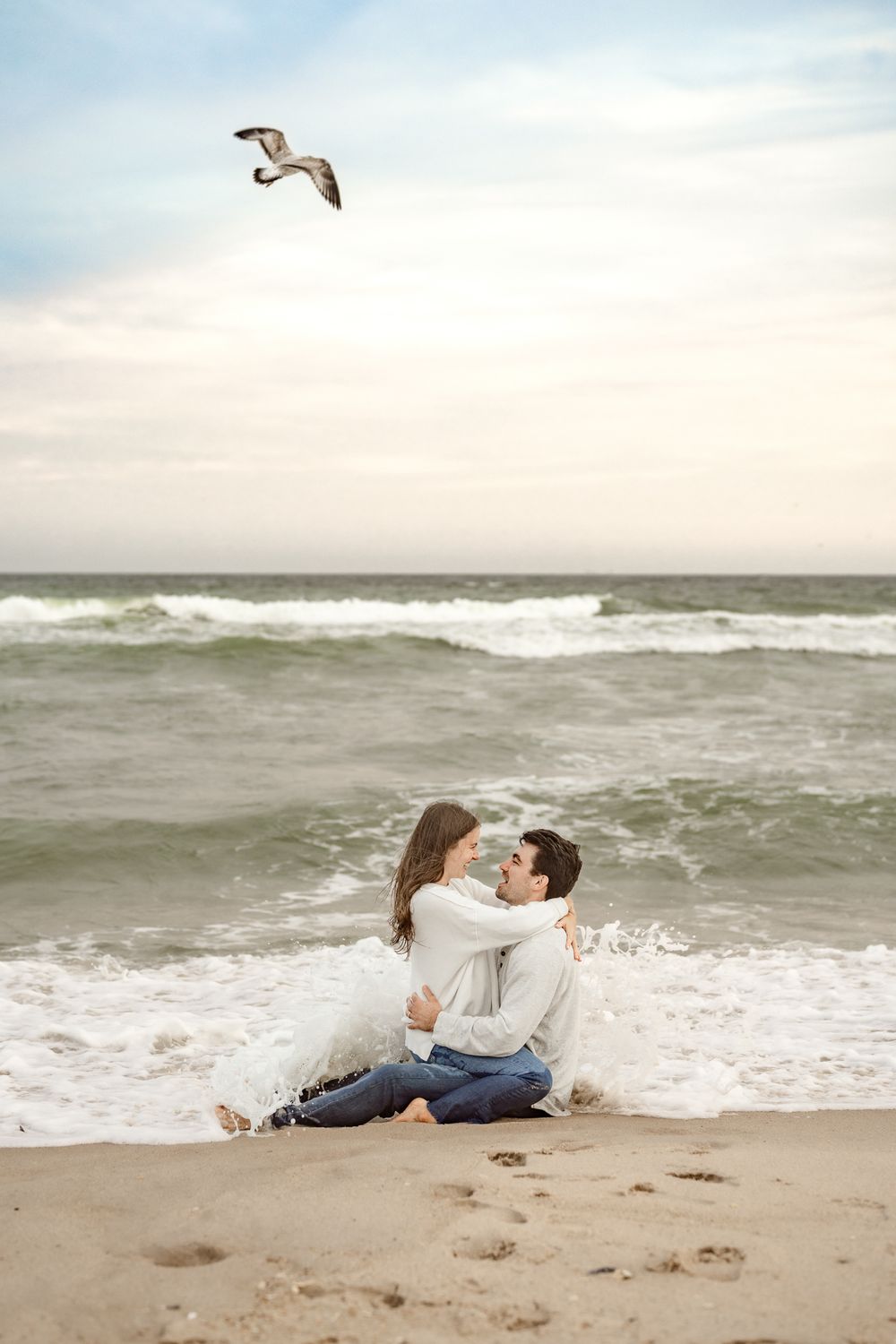 A romantic couple plays in the ocean waves on a cloudy beach day as a seagull flies overhead.